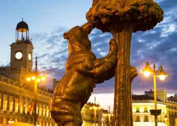 Statue of bear and madrone tree, famous symbol of city of Madrid on busy Puerta del Sol square at dusk, Spain. Square composition.